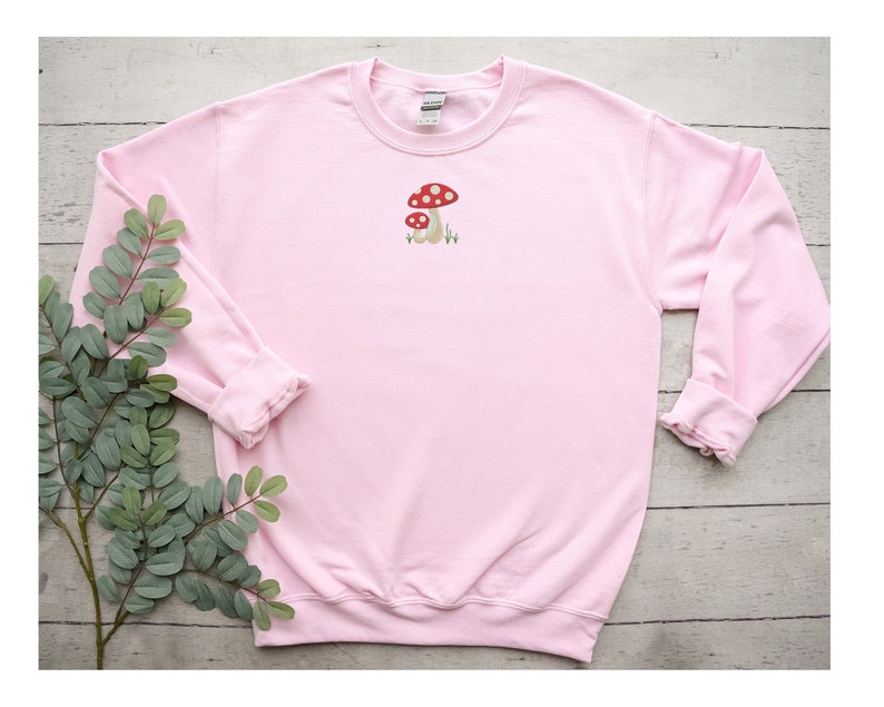 Pode incluir: Uma camisola cor-de-rosa com uma pequena imagem bordada de dois cogumelos vermelhos e brancos na frente.