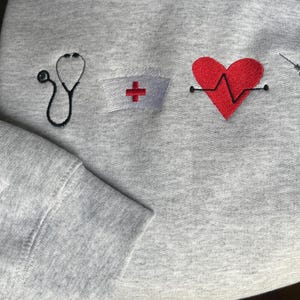 May include: Gray sweatshirt with red embroidered medical symbols, including a stethoscope, a heart with a heartbeat line, a syringe, and a nurse's cap with a red cross.