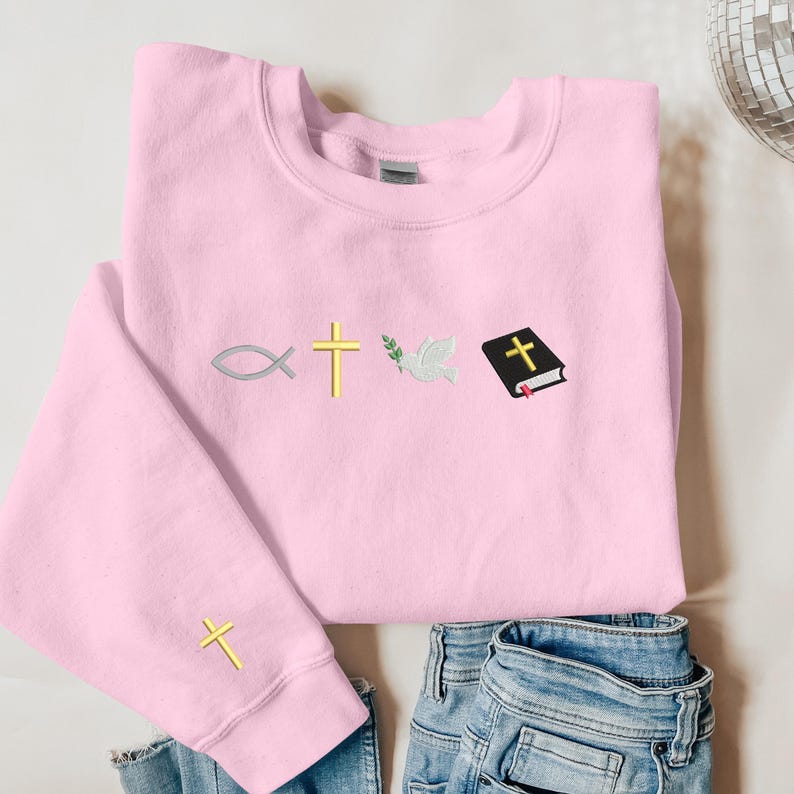 May include: A pink sweatshirt with a cross, a dove, a fish, and a closed book with a cross on it embroidered on the front. The sweatshirt has a small cross embroidered on the left sleeve.