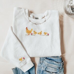 K&ouml;nnte beinhalten: Wei&szlig;er Pullover mit einem gestickten Design aus gelbem Huhn und K&uuml;ken. Der Pullover hat einen Rundhalsausschnitt und lange &Auml;rmel.