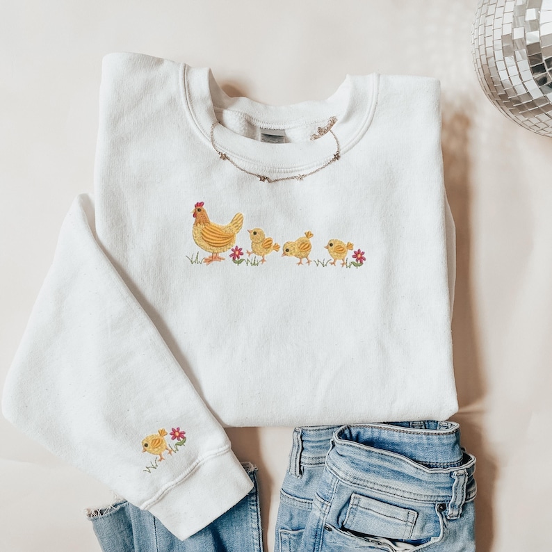 K&ouml;nnte beinhalten: Wei&szlig;er Pullover mit einem gestickten Design aus gelbem Huhn und K&uuml;ken. Der Pullover hat einen Rundhalsausschnitt und lange &Auml;rmel.