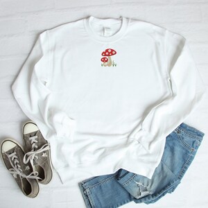 Pode incluir: Camisola branca com um gr&aacute;fico de cogumelo vermelho e branco bordado na frente. A camisola &eacute; combinada com cal&ccedil;as de ganga azuis e t&eacute;nis cinzentos.