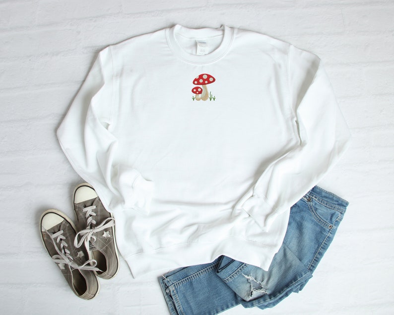 Pode incluir: Camisola branca com um gr&aacute;fico de cogumelo vermelho e branco bordado na frente. A camisola &eacute; combinada com cal&ccedil;as de ganga azuis e t&eacute;nis cinzentos.