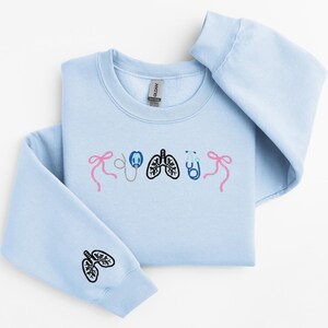 May include: Light blue sweatshirt with embroidered medical-themed designs. Features pink bows, a stethoscope, lungs, and a stethoscope. The left sleeve has a lung design. A comfortable, casual top for medical professionals or students.