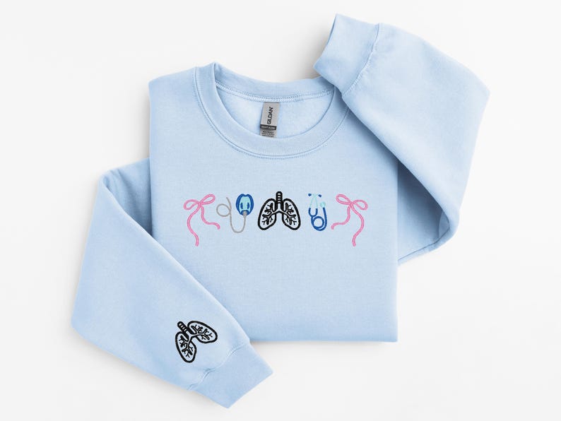 May include: Light blue sweatshirt with embroidered medical-themed designs. Features pink bows, a stethoscope, lungs, and a stethoscope. The left sleeve has a lung design. A comfortable, casual top for medical professionals or students.