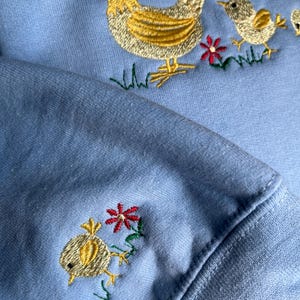 K&ouml;nnte beinhalten: Ein hellblauer Pullover mit gestickten gelben K&uuml;ken und einer Henne mit roten Akzenten. Die Henne ist von gr&uuml;nem Gras und roten Blumen umgeben.