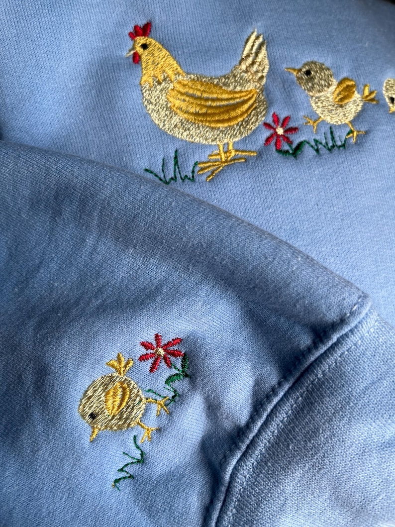 K&ouml;nnte beinhalten: Ein hellblauer Pullover mit gestickten gelben K&uuml;ken und einer Henne mit roten Akzenten. Die Henne ist von gr&uuml;nem Gras und roten Blumen umgeben.