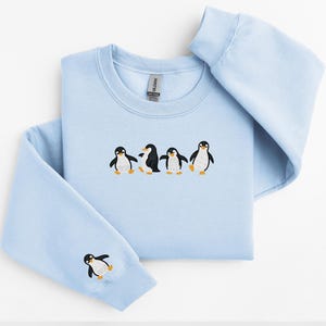 Pu&ograve; includere: Felpa azzurra con ricamo di pinguini. La felpa girocollo presenta una fila di cinque pinguini ricamati sul petto, con un singolo pinguino sulla manica sinistra. I pinguini sono neri, bianchi e arancioni.
