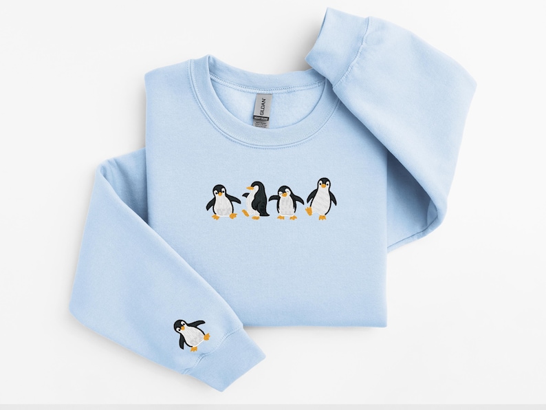 Pu&ograve; includere: Felpa azzurra con ricamo di pinguini. La felpa girocollo presenta una fila di cinque pinguini ricamati sul petto, con un singolo pinguino sulla manica sinistra. I pinguini sono neri, bianchi e arancioni.