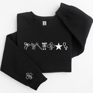 May include: Black crewneck sweatshirt with white medical-themed graphics. The design includes a bow, knee joint, hammer, medical symbol, cross, star, and stethoscope. A smaller cross graphic is on the sleeve cuff.
