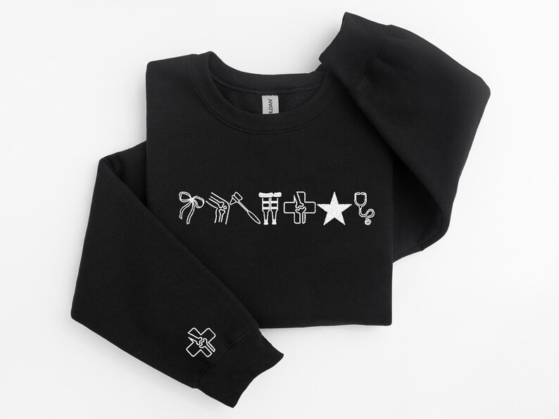 May include: Black crewneck sweatshirt with white medical-themed graphics. The design includes a bow, knee joint, hammer, medical symbol, cross, star, and stethoscope. A smaller cross graphic is on the sleeve cuff.