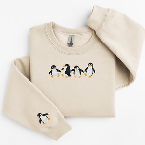 Pu&ograve; includere: Una felpa beige con girocollo e maniche lunghe. La parte anteriore della felpa presenta un disegno ricamato di quattro pinguini. Un pinguino &egrave; ricamato anche sulla manica sinistra.