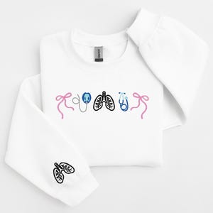 May include: White sweatshirt with embroidered medical-themed graphics. The design includes a pink bow, an oxygen mask, lungs, a stethoscope, and a blue bow. A lung graphic is also on the sleeve. A comfortable, casual top for medical professionals.