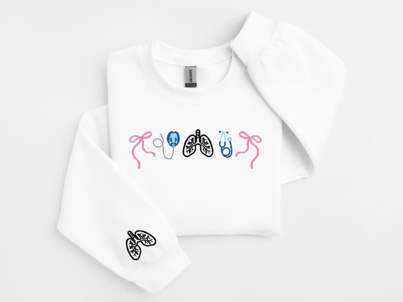 May include: White sweatshirt with embroidered medical-themed graphics. The design includes a pink bow, an oxygen mask, lungs, a stethoscope, and a blue bow. A lung graphic is also on the sleeve. A comfortable, casual top for medical professionals.