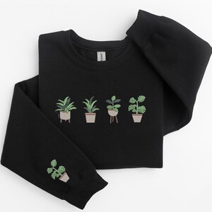 Pode incluir: Camisola de moletom preta com um design bordado verde de plantas em vasos. O design apresenta cinco plantas em vasos, com tr&ecirc;s na frente e duas nas mangas.