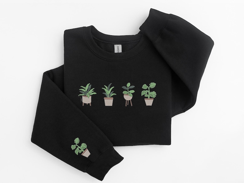Pode incluir: Camisola de moletom preta com um design bordado verde de plantas em vasos. O design apresenta cinco plantas em vasos, com tr&ecirc;s na frente e duas nas mangas.