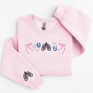 May include: A light pink sweatshirt with embroidered medical-themed designs. The design includes a stethoscope, lungs, and bows. The left sleeve features a lung design. The sweatshirt is made of a soft material.