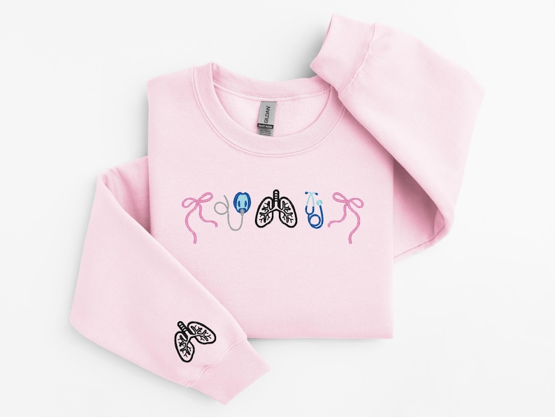 May include: A light pink sweatshirt with embroidered medical-themed designs. The design includes a stethoscope, lungs, and bows. The left sleeve features a lung design. The sweatshirt is made of a soft material.