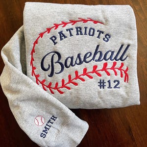 May include: A gray sweatshirt with the words "PATRIOTS Baseball #12" embroidered in navy blue and red. The sleeve features a baseball and the name "SMITH" embroidered in navy blue. The design is centered on the chest.