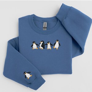 Pu&ograve; includere: Una felpa blu con pinguini ricamati. La felpa girocollo presenta una fila di quattro pinguini sul petto e un singolo pinguino sulla manica sinistra. I pinguini sono neri, bianchi e gialli.