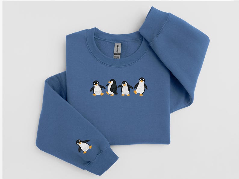 Pu&ograve; includere: Una felpa blu con pinguini ricamati. La felpa girocollo presenta una fila di quattro pinguini sul petto e un singolo pinguino sulla manica sinistra. I pinguini sono neri, bianchi e gialli.