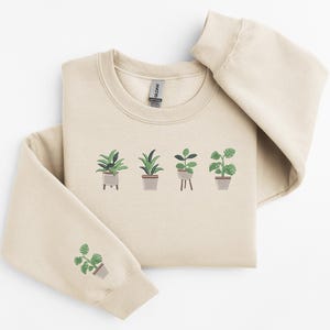 Pode incluir: Uma camisola bege com um gr&aacute;fico de cinco plantas em vasos na frente. A camisola tem um decote redondo e mangas compridas.