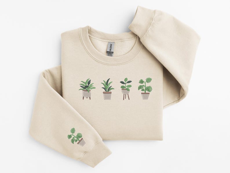 Pode incluir: Uma camisola bege com um gr&aacute;fico de cinco plantas em vasos na frente. A camisola tem um decote redondo e mangas compridas.