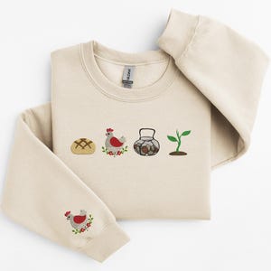 May include: A beige sweatshirt with an embroidered design of a loaf of bread, a chicken, a basket of eggs, and a plant. The sweatshirt has a crew neck and long sleeves.