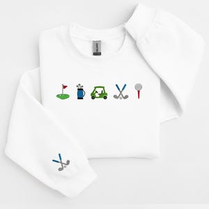 以下が含まれることがあります： ゴルフをテーマにした刺繍が施された白いスウェットシャツ。デザインには、ゴルフの旗、ゴルフバッグ、ゴルフカート、ゴルフクラブ、ゴルフボールが含まれています。