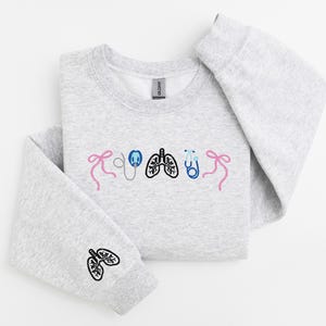 May include: A light gray sweatshirt with embroidered medical-themed designs. The design includes pink bows, a stethoscope, lungs, and a stethoscope. The left sleeve features a lung design. The sweatshirt is folded on a white surface.