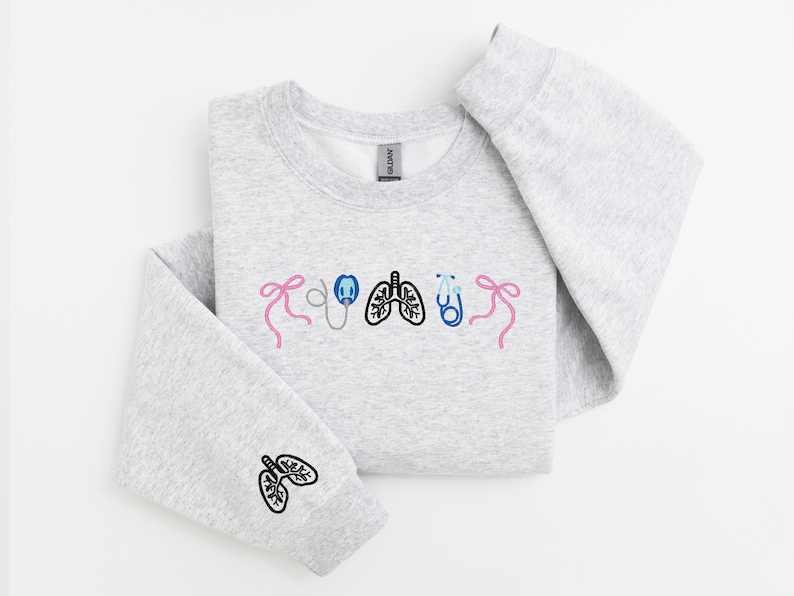 May include: A light gray sweatshirt with embroidered medical-themed designs. The design includes pink bows, a stethoscope, lungs, and a stethoscope. The left sleeve features a lung design. The sweatshirt is folded on a white surface.