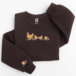 K&ouml;nnte beinhalten: Ein dunkelbraunes Sweatshirt mit einem gestickten Design einer Henne mit ihren K&uuml;ken. Die K&uuml;ken sind gelb und die Henne ist braun mit einem roten Kamm.