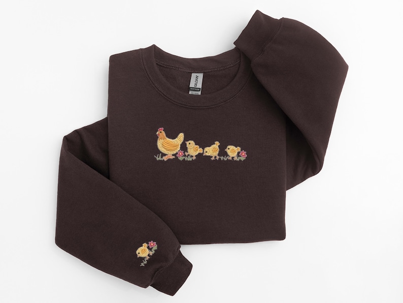 K&ouml;nnte beinhalten: Ein dunkelbraunes Sweatshirt mit einem gestickten Design einer Henne mit ihren K&uuml;ken. Die K&uuml;ken sind gelb und die Henne ist braun mit einem roten Kamm.