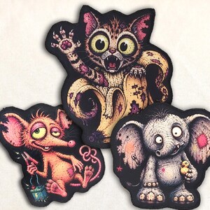 Könnte beinhalten: Drei gestickte Aufnäher mit stilisierten Cartoon-Kreaturen: eine Katze, die aus einer Bananenschale kommt, eine rosa Maus, die Starthilfekabel an einer Batterie hält, und ein grauer Elefant, der ein kleines gelbes Entenküken hält, alle mit schwarzem Rand.