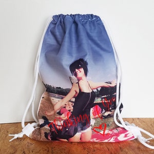 May include: A white drawstring bag with a photo printed on the front. The photo shows a woman wearing sunglasses and a black tank top, smiling and holding her arms out. The text "Thinking of you" is printed in red on the bag.