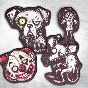 Può includere: Quattro toppe ricamate con disegni scuri e illustrativi: un cane con un occhio verde neon, un volto di clown angosciato con accenti rossi, un topo malandato e una piccola figura di angelo con aureola, in nero, bianco e rosso.