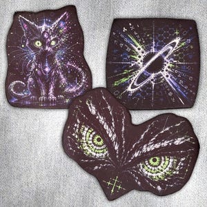 Könnte beinhalten: Drei Stoffaufnäher mit dunklen, komplexen Motiven. Ein Aufnäher zeigt eine Katze mit leuchtend grünen Augen und violetten kosmischen Mustern. Ein weiterer ist quadratisch mit einem weißen Planetenring und geometrischen Linien. Der dritte zeigt zwei große grüne Augen.