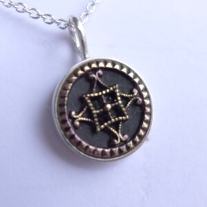 Op de afbeelding: Een zilveren ketting met een ronde hanger. De hanger is zwart met een goudkleurig ontwerp in het midden.