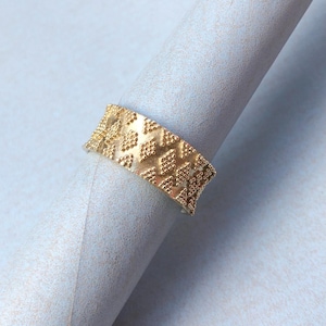 Può includere: Un anello color oro con una superficie strutturata con un motivo ripetuto di piccole forme in rilievo. L'anello è esposto su una superficie arrotolata grigio chiaro, su uno sfondo azzurro. Il design dell'anello è visibile.