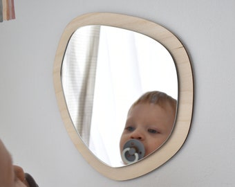 May include: A wooden mirror with a unique organic shape, featuring a rounded edge and a smooth, light wood finish. A baby is looking at their reflection in the mirror.