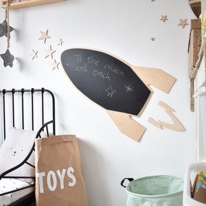 May include: A white wall with a wooden rocket ship chalkboard with the words "To the moon and back" written on it. There are several wooden stars on the wall. A brown paper bag with the word "TOYS" printed on it is on the floor. A green fabric storage bin with a black bear face on it is on the floor.