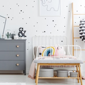 Könnte beinhalten: Ein weißes Metallbettgestell mit einem grauen, gepolsterten Kopfteil und Fußteil. Das Bett ist mit einem Regenbogenkissen und einem rosa Kissen dekoriert. Die Wand hinter dem Bett ist mit schwarzen Stern-Stickern verziert. Im Hintergrund befindet sich eine graue Kommode mit goldenen Kronen-Schubladenknöpfen.