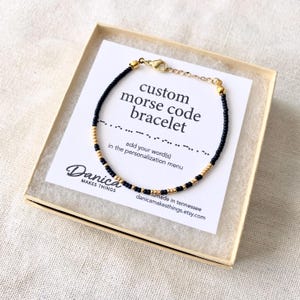 May include: A black and gold beaded bracelet with a custom morse code message. The bracelet is in a small box with a white card that says "custom morse code bracelet" and "add your word(s) in the personalization menu".