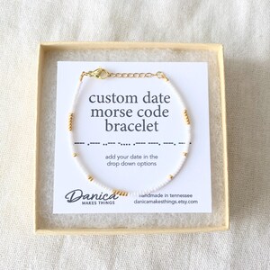 May include: White beaded bracelet with gold beads spelling out a date in Morse code. The bracelet is in a box with a card that says "custom date morse code bracelet" and "add your date in the drop down options". The card also has the text "Danica Makes Things" and "handmade in tennessee danicamakesthings.etsy.com".