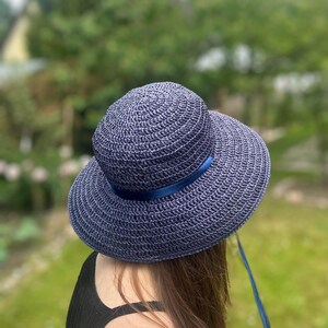 Op de afbeelding: Een marineblauwe zonnehoed met een brede rand en een bijpassende lintband. De hoed is gemaakt van geweven materiaal en heeft een afgeronde kroon. Een blauw lint is om de basis van de kroon gebonden.