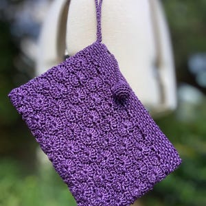 May include: A handcrafted, purple crochet clutch with a textured floral pattern. The clutch features a small, round button closure and a matching loop handle. The bag is made of a shiny, possibly raffia-like material.