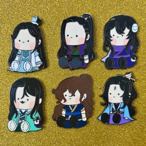 Op de afbeelding: Zes cartoonpersonagespinnen met zwart haar en verschillende outfits. De pinnen tonen personages in tinten blauw, paars, groen en bruin. Elk personage zit met gekruiste benen. De achtergrond is een goudkleurig glitteroppervlak.