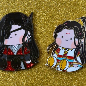 May include: Two enamel pins featuring chibi-style characters with black hair and detailed outfits. One pin has a red and black outfit with an eye patch, the other has a white and gold outfit. Both pins have a gold outline and are set against a gold glitter background.