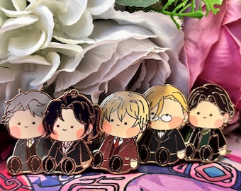 May include: Five gold-colored enamel pins featuring cartoon characters wearing suits. The characters have different hair colors and styles. The pins are set against a background of white and pink flowers.