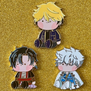 May include: Three enamel pins featuring chibi-style anime characters with large heads and seated poses. The pins have gold outlines and are set against a gold glitter background. Each character has unique hair and clothing.